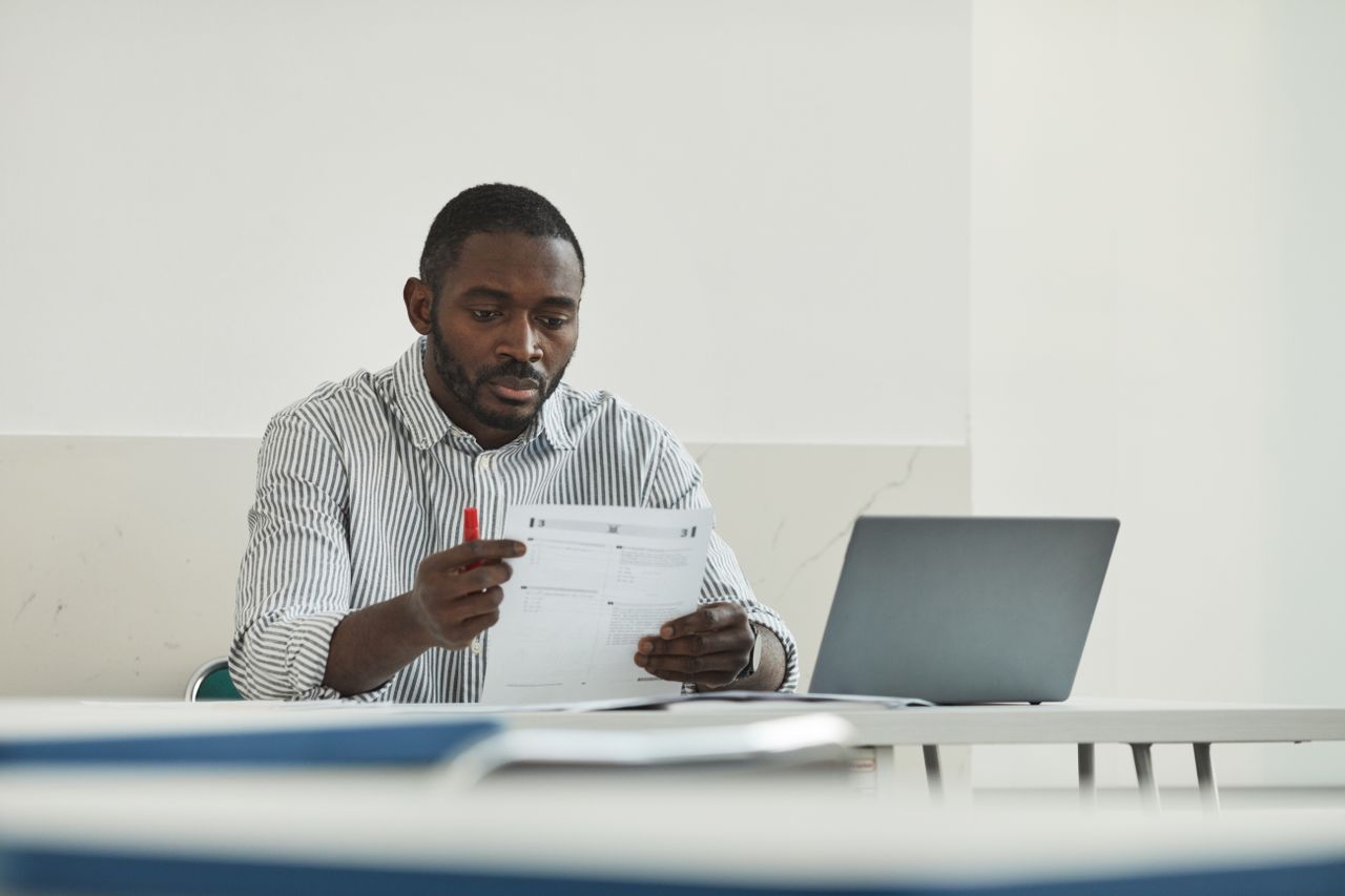 An educator reviewing assessment sheets at his desk next to an open laptop