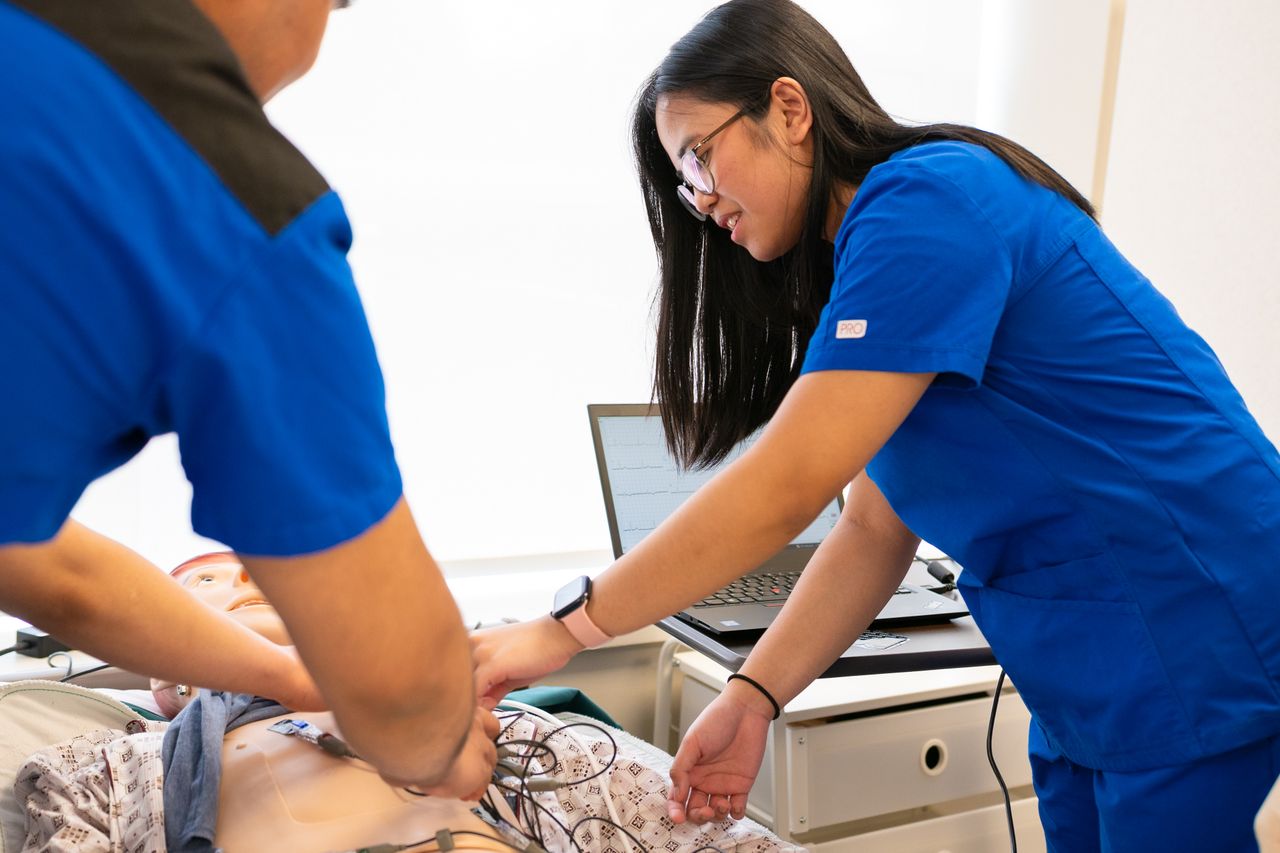 Nursing students practicing on a patient manikin in a simulation lab, with vital signs on a laptop screen