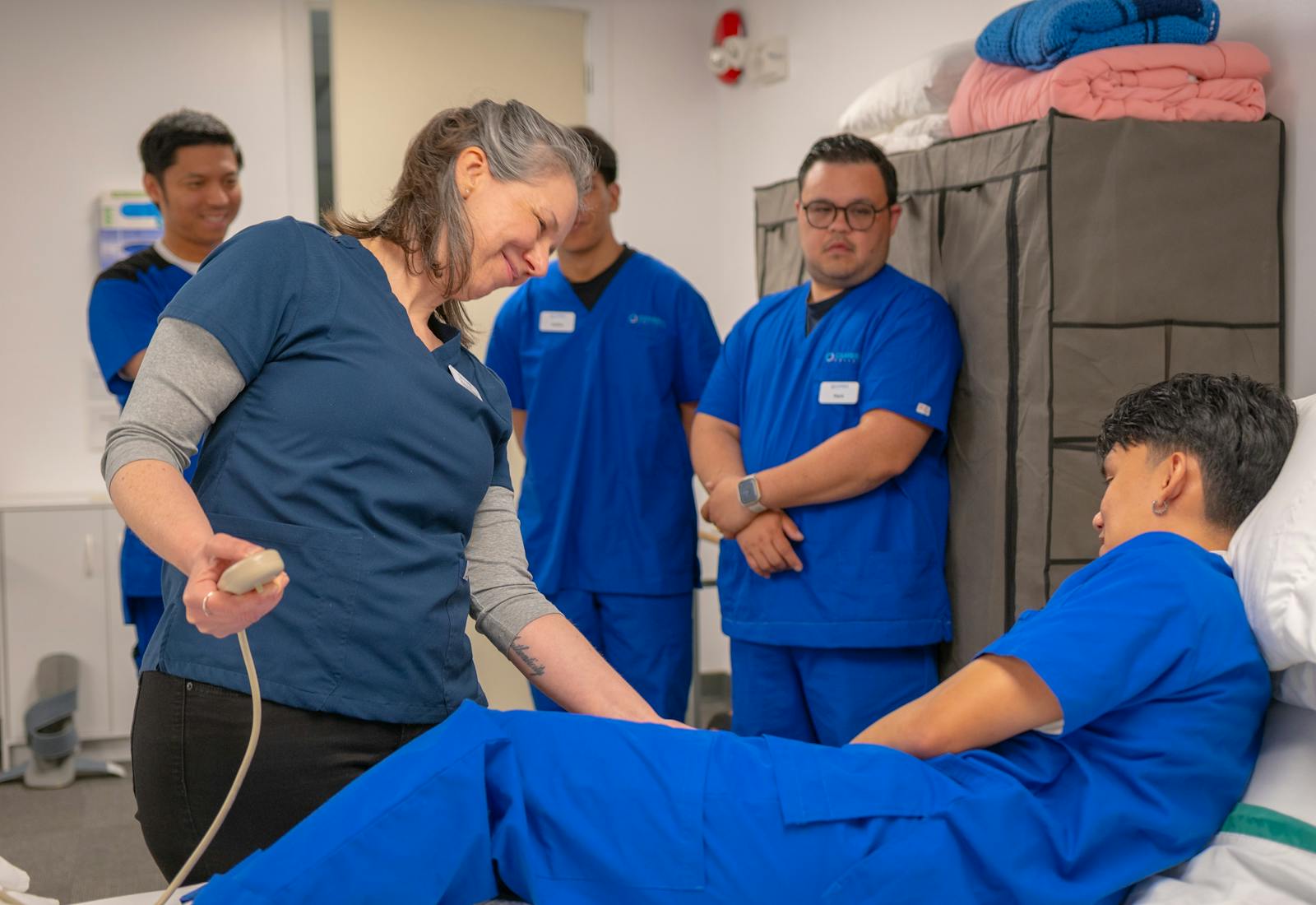 A nursing instructor guiding a group of CNA students around a practice bed.