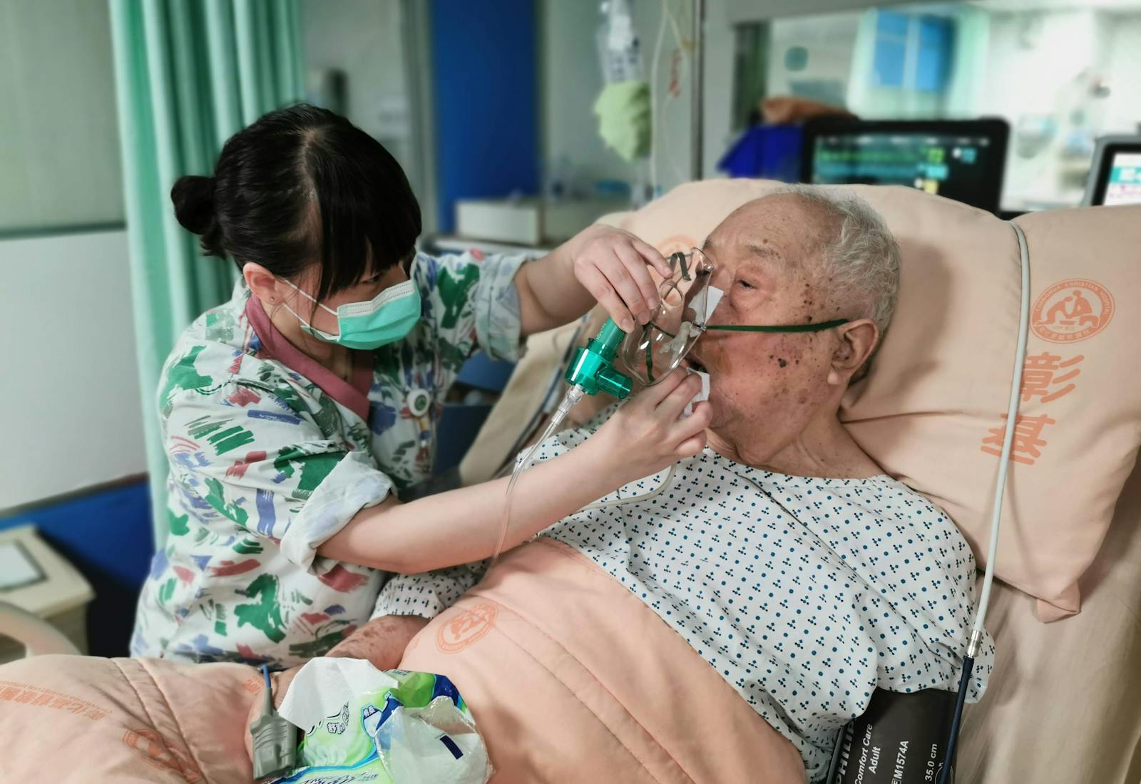 A caregiver assisting an elderly patient with an oxygen mask in a hospital bed.